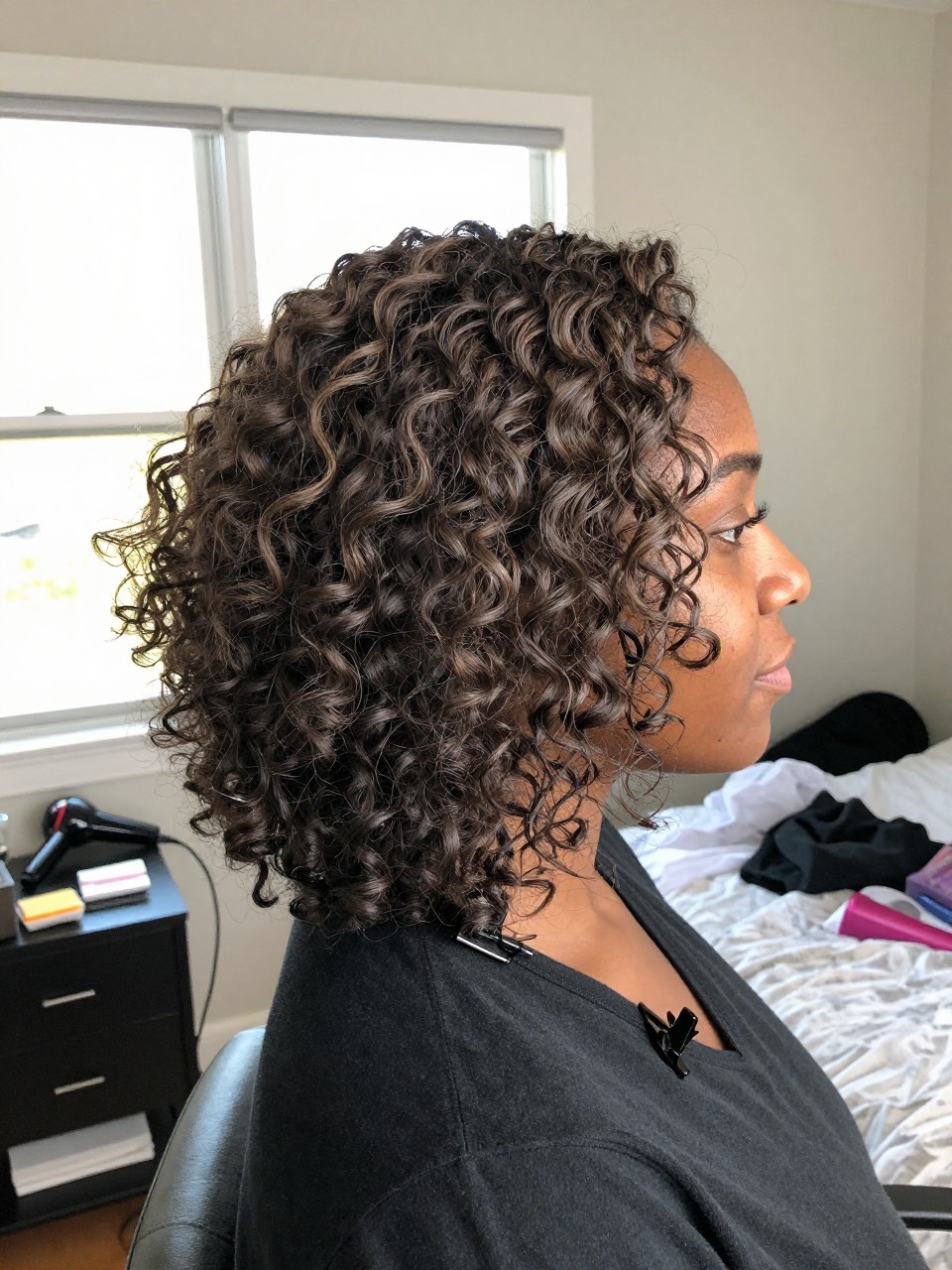 Photo of a Black woman wearing stretched natural curls with ash brown highlights softly blended throughout, side profile view, in a sun-drenched bedroom, natural window light, containing a small lived-in detail such as a blow dryer on the counter and a claw clip clipped to the collar, latest iPhone photo quality.