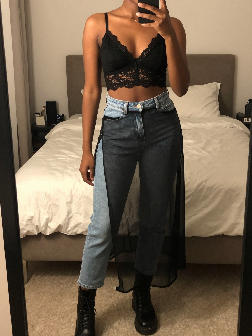 mirror selfie of a black woman wearing a black lace bralette with light wash mom jeans underneath a black sheer mesh skirt and combat boots, one hand holding her phone at chest level, cropped at NECK so face doesn't show, in a feminine-themed bedroom, warm moody lighting, iPhone photo quality.