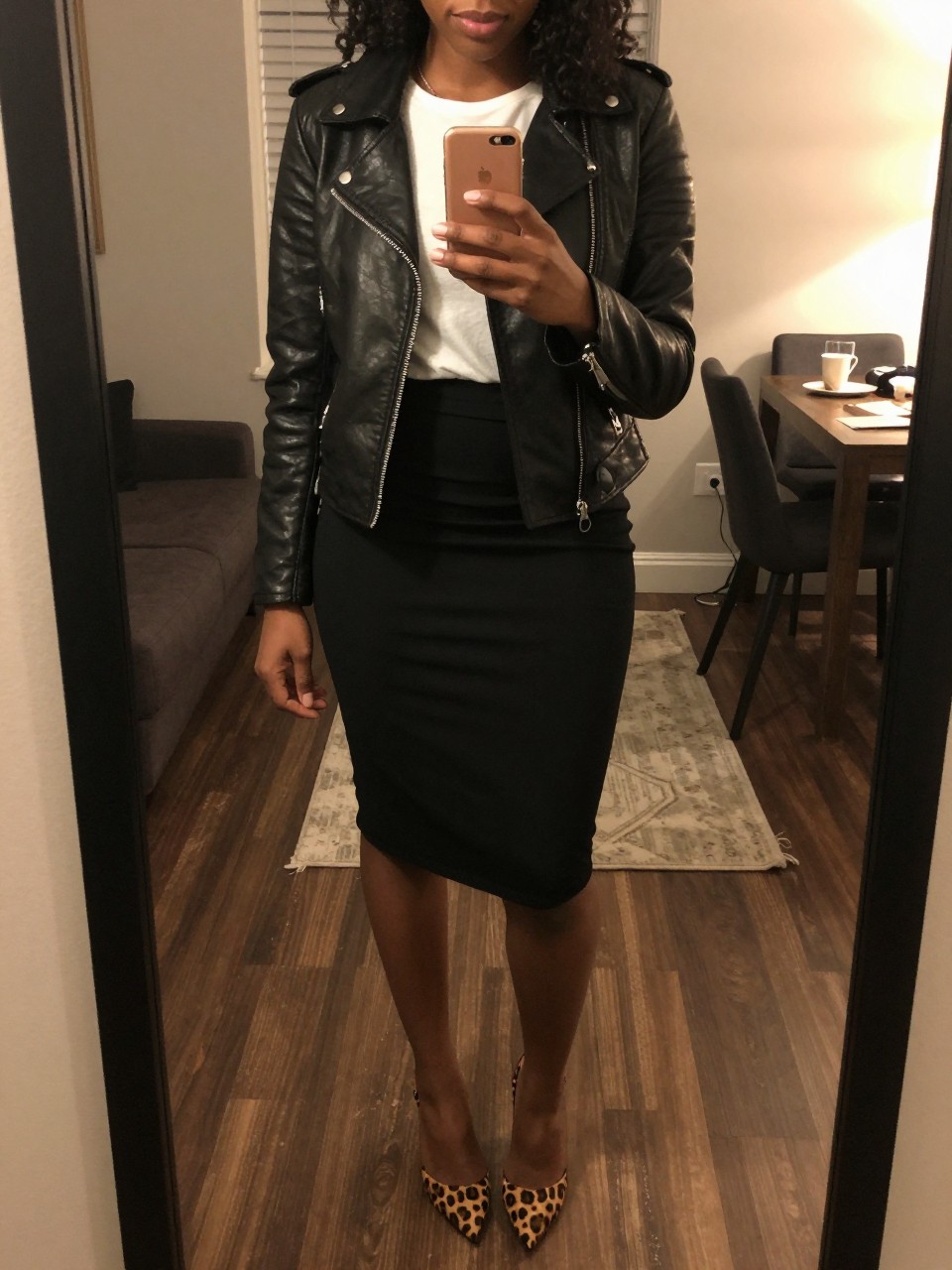 mirror selfie of a black woman wearing a black leather jacket over a white tee and black pencil skirt with leopard print heels, holding her phone at chest level, cropped at NECK so face doesn't show, in a city apartment, warm moody lighting, iPhone photo quality.