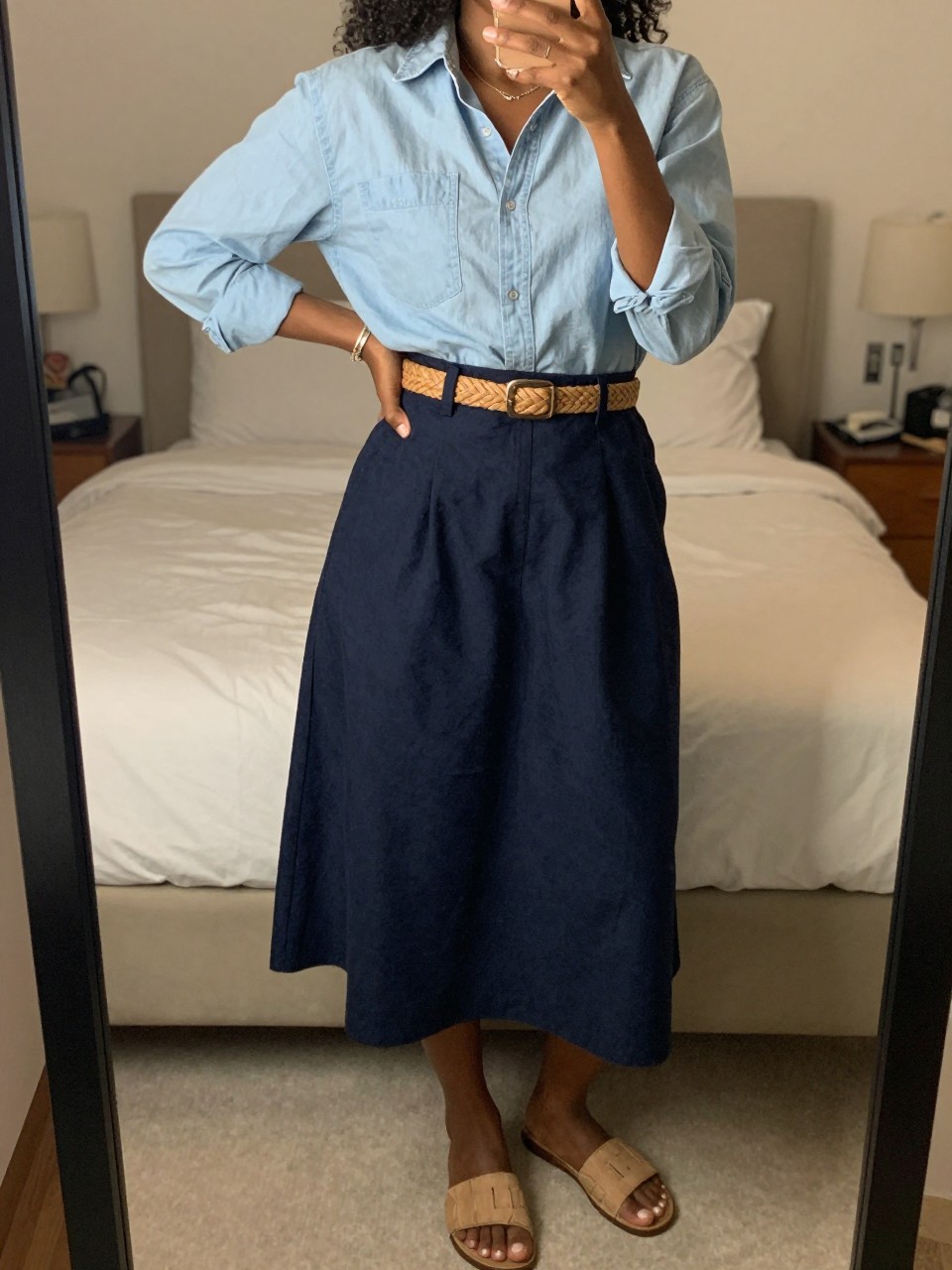 iPhone mirror selfie of a black woman wearing a light blue chambray shirt tucked into a navy cotton A-line skirt with a woven tan belt and slide sandals, one hand on her hip, cropped at NECK so face doesn't show, in a feminine-themed bedroom, warm moody lighting, iPhone photo quality.