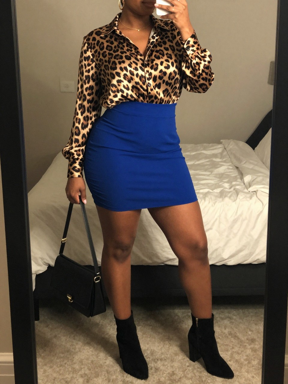 mirror selfie of a curvy black woman wearing a leopard print silk blouse tucked into a cobalt blue mini skirt with black suede block-heel ankle boots and gold hoop earrings, holding a black crossbody bag, standing in a confident pose, cropped at NECK so face doesn't show, in a feminine-themed bedroom, warm moody lighting, iPhone photo quality.