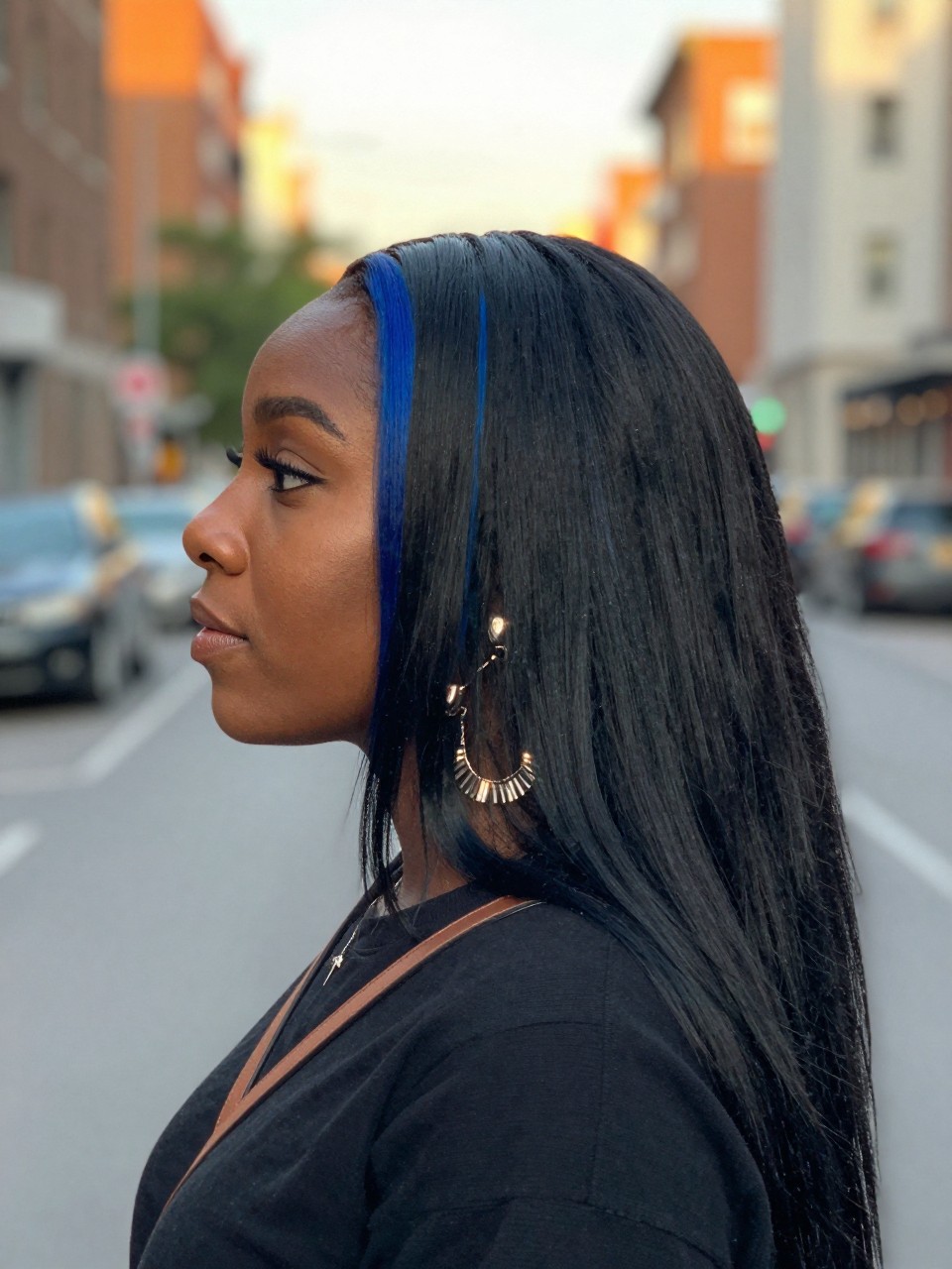 Photo of a black woman wearing long straight black hair with blue face-framing highlights, side profile view, in a softly blurred city street background, golden hour glow, containing a small lived-in detail such as statement earrings peeking through and a crossbody strap on the shoulder, latest iPhone photo quality.