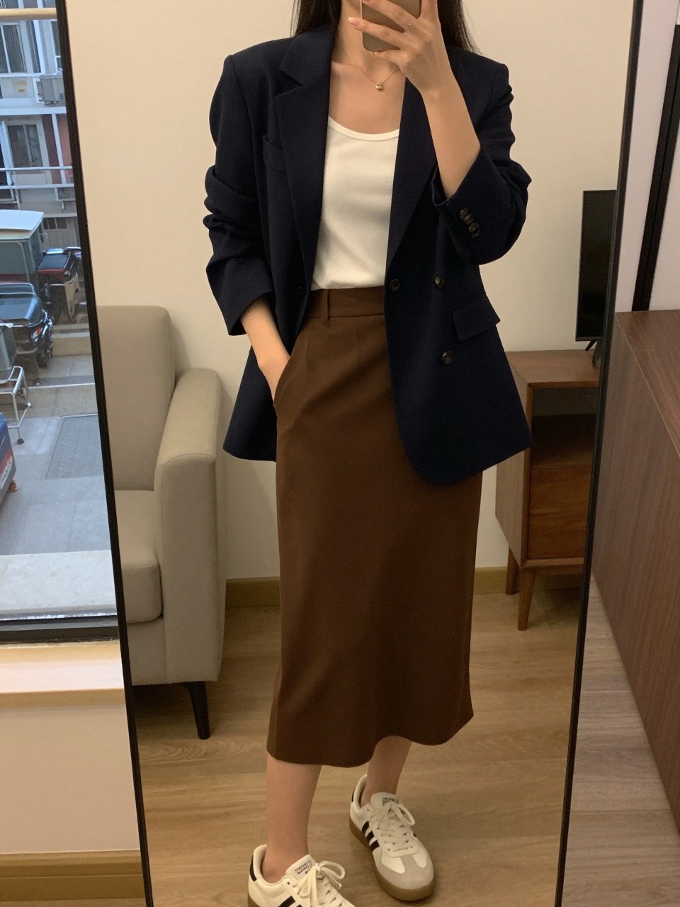 iPhone mirror selfie of a woman wearing a navy blazer over a white tank top tucked into a chocolate brown midi skirt with white sneakers, one hand in her blazer pocket, cropped at NECK so face doesn't show, in a city apartment, warm moody lighting, iPhone photo quality.
