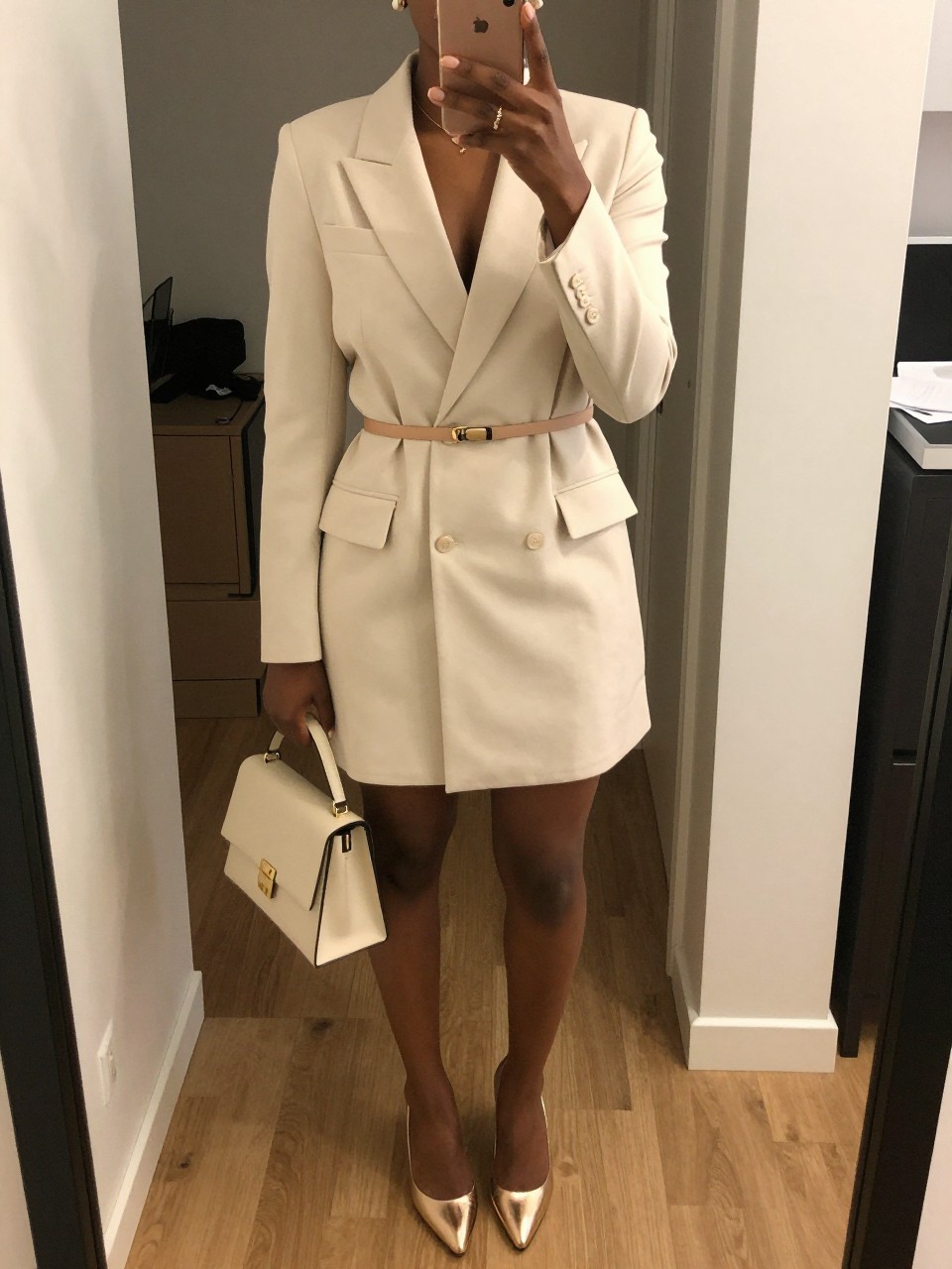 mirror selfie of a black woman wearing a cream tailored blazer dress with a thin nude waist belt, champagne gold metallic heels, a cream mini top-handle bag, and pearl stud earrings, holding a phone in front of a mirror, cropped at NECK so face doesn't show, with a natural casual selfie pose, in a city apartment, warm moody lighting, including small lived-in details latest iPhone photo quality.