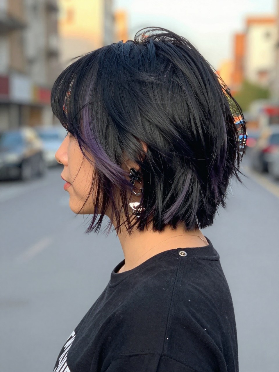 Photo of a woman wearing a tousled black bob with lavender highlights and soft choppy texture, back-of-head view to highlight details, in a softly blurred city street background, golden hour glow, containing a small lived-in detail such as a hair clip clipped to the collar and statement earrings peeking through, latest iPhone photo quality.