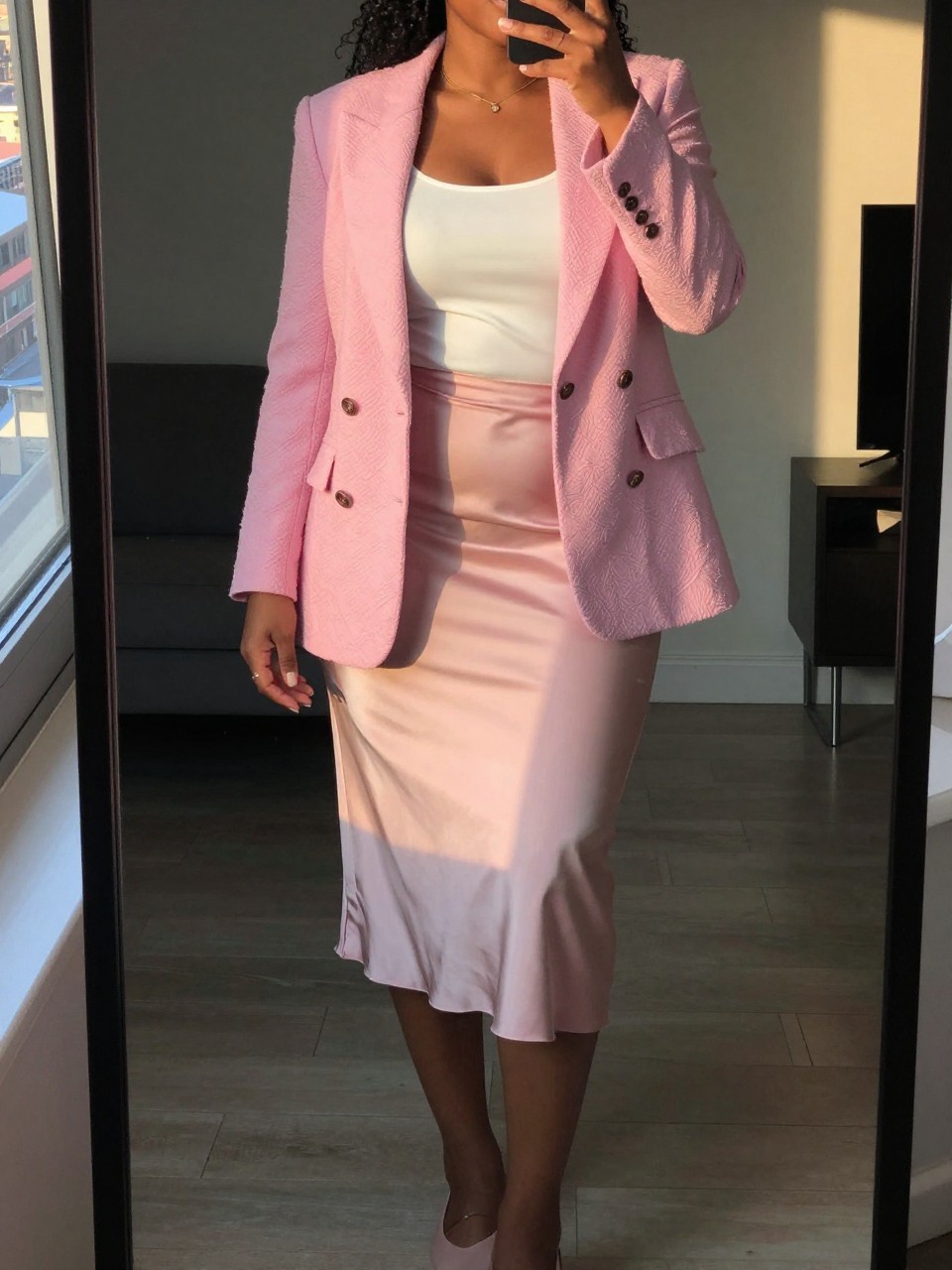 mirror selfie of a curvy black woman in a textured pink blazer and white top with a satin midi skirt and flats, standing with slight hip tilt, cropped at NECK so face doesn't show, in a city apartment, evening golden hour, iPhone photo quality.