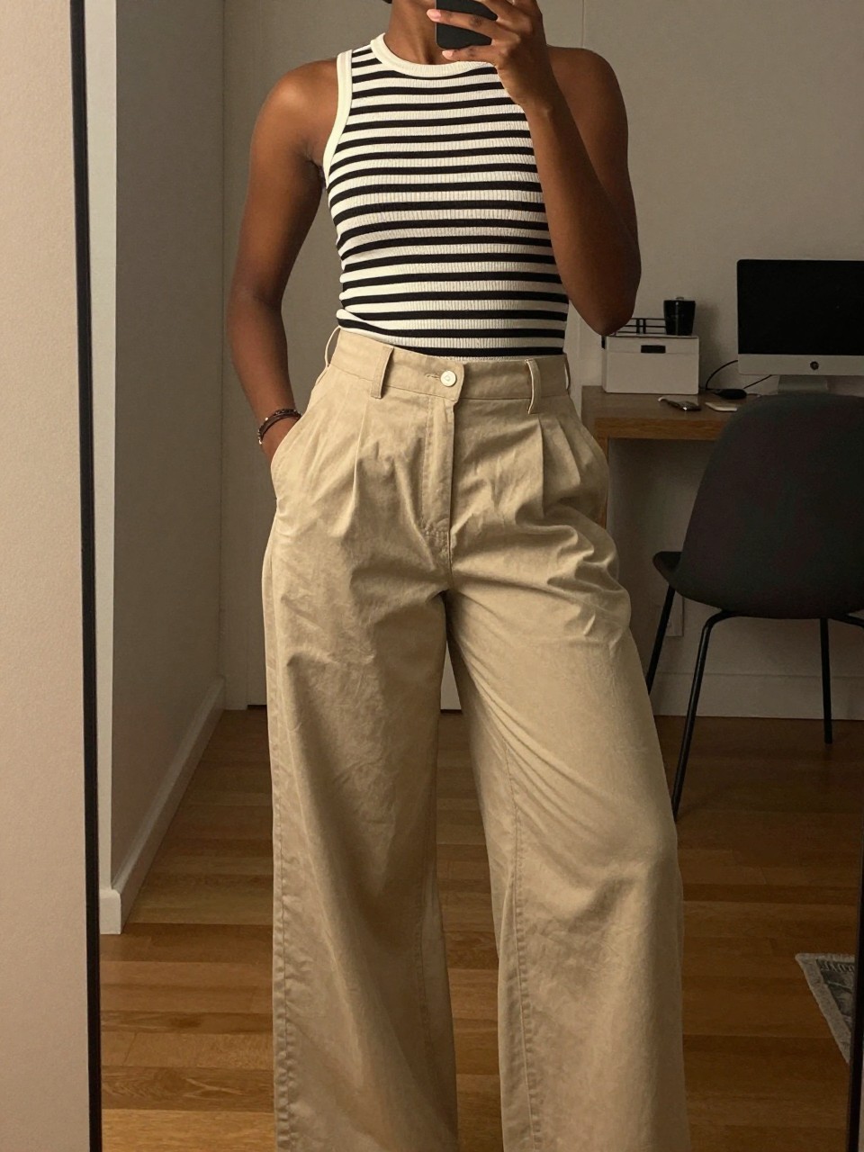 mirror selfie of a black woman in a striped top and wide-leg pants, posing hands in pockets, cropped at NECK so face doesn't show, in a city apartment, warm moody lighting, iPhone photo quality.