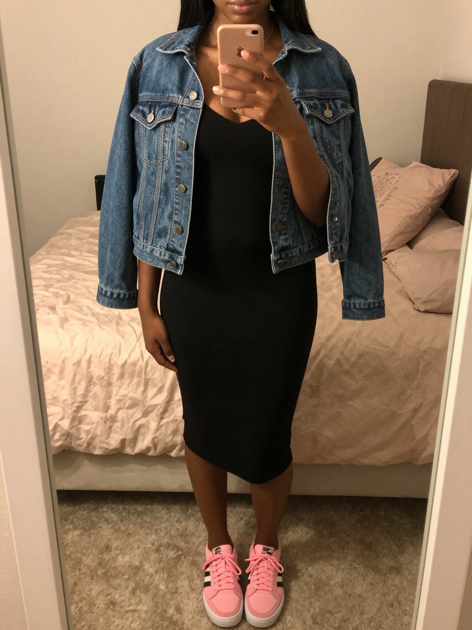 iPhone mirror selfie of a black woman in a black slip dress and denim jacket with pink sneakers, holding phone at chest level, cropped at NECK so face doesn't show, in a feminine-themed bedroom, warm moody lighting, iPhone photo quality.