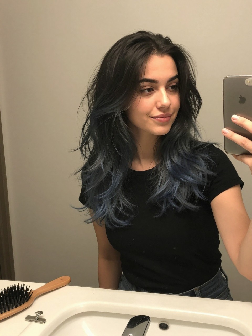 Photo of a woman wearing a wavy black lob with dusty blue highlights, candid mirror selfie, at a bathroom vanity mirror, soft indoor lighting, containing a small lived-in detail such as a hairbrush on the sink and a silver clip nearby, latest iPhone photo quality.