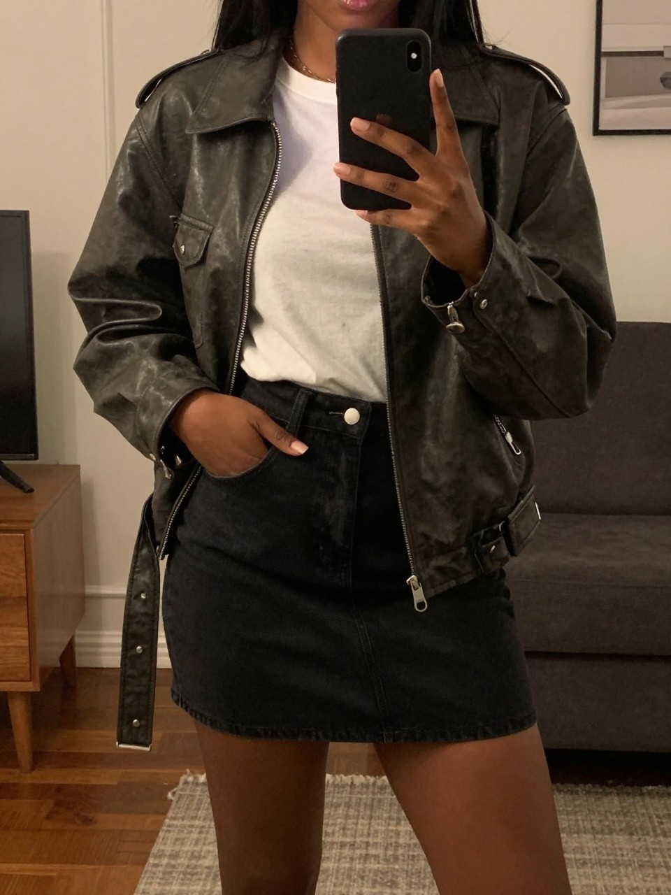mirror selfie of a Black woman wearing a black denim skirt with a leather jacket and white tee, standing with one hand in her jacket pocket and phone in the other, cropped at NECK so face doesn't show, in a city apartment, warm moody lighting, iPhone photo quality.
