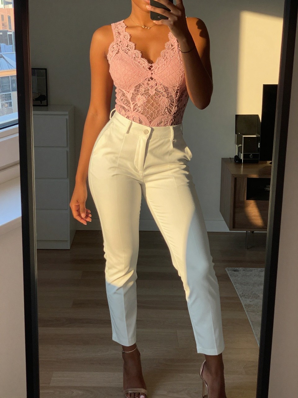 mirror selfie of a black woman in a pink lace top with white trousers and heels, standing with slight hip tilt, cropped at NECK so face doesn't show, in a city apartment, evening golden hour, iPhone photo quality.