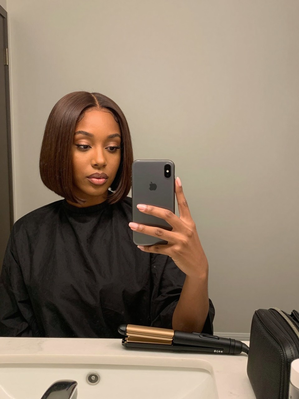 Photo of a black woman wearing a mahogany brown bob styled sleek with softly curved ends, candid mirror selfie, bathroom vanity mirror setting, soft indoor lighting, with a flat iron on the counter and a small makeup bag nearby, latest iPhone photo quality.