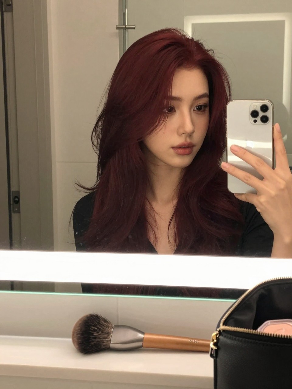 Photo of a woman wearing mahogany red hair in a soft bouncy blowout, candid mirror selfie, bathroom vanity mirror setting, soft indoor lighting, with a round brush on the counter and a makeup bag nearby, latest iPhone photo quality.