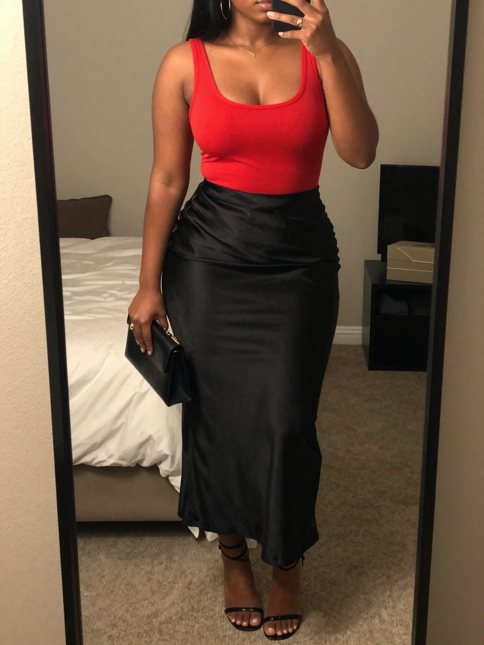 iPhone mirror selfie of a curvy black woman wearing a fitted true red scoop-neck top tucked into a black satin maxi skirt with black strappy heeled sandals and minimal gold hoops, holding a small black clutch, standing in a confident color-block pose, cropped at NECK so face doesn't show, in a feminine-themed bedroom, warm moody lighting, iPhone photo quality.