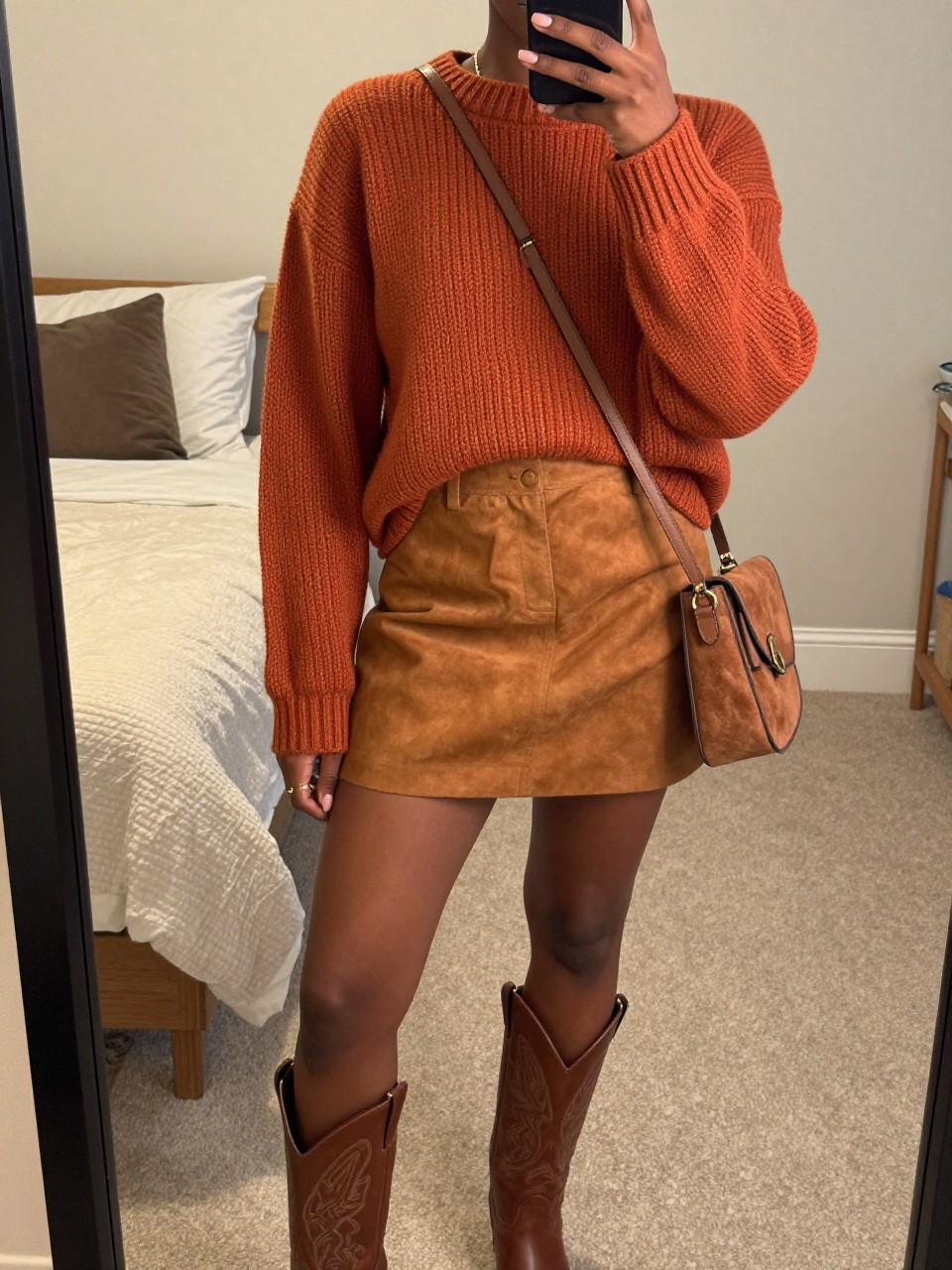 mirror selfie of a black woman wearing a rust orange chunky sweater tucked into a caramel brown suede skirt with brown leather western boots and a matching suede crossbody bag, sunglasses perched on her head, standing in a relaxed pose, cropped at NECK so face doesn't show, in a feminine-themed bedroom, warm moody lighting, iPhone photo quality.