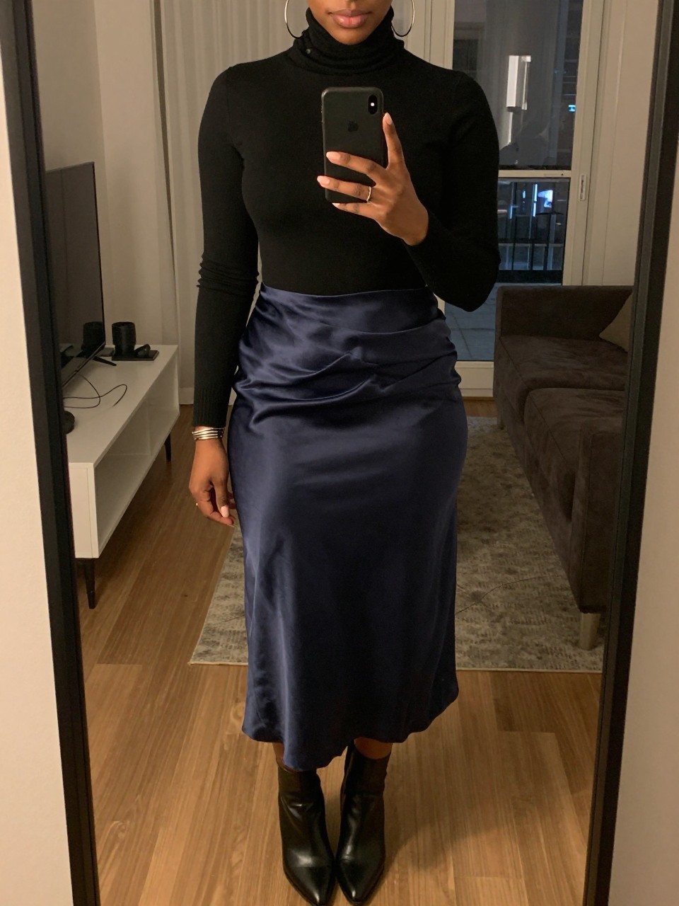 iPhone mirror selfie of a black woman wearing a black turtleneck tucked into a navy satin midi skirt with large silver hoops and black ankle boots, holding her phone at chest level, cropped at NECK so face doesn't show, in a city apartment, warm moody lighting, iPhone photo quality.