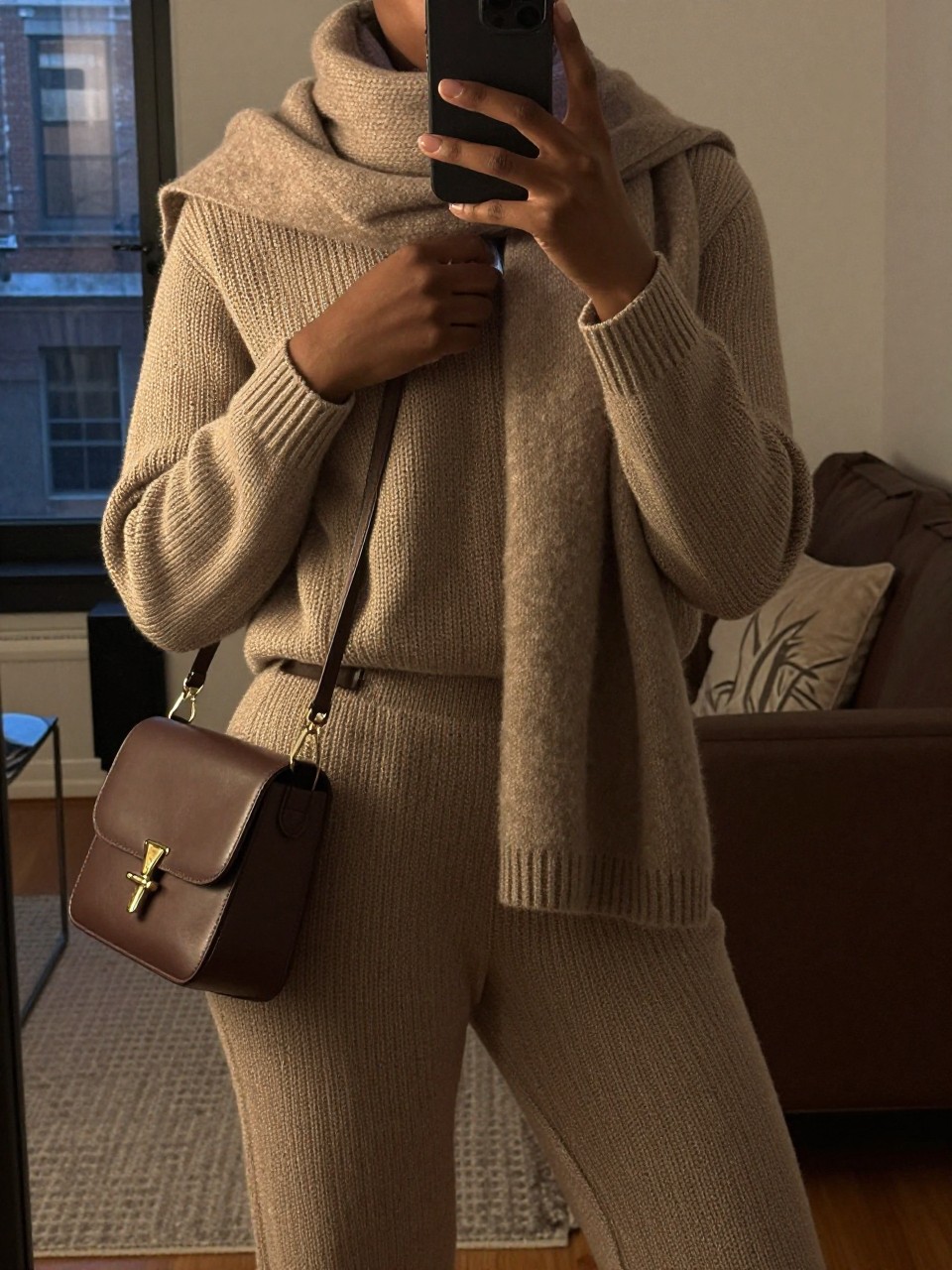 iPhone mirror selfie of a black woman in a knit set, adjusting the scarf on her bag, cropped at NECK so face doesn't show, in a city apartment, warm moody lighting, iPhone photo quality.