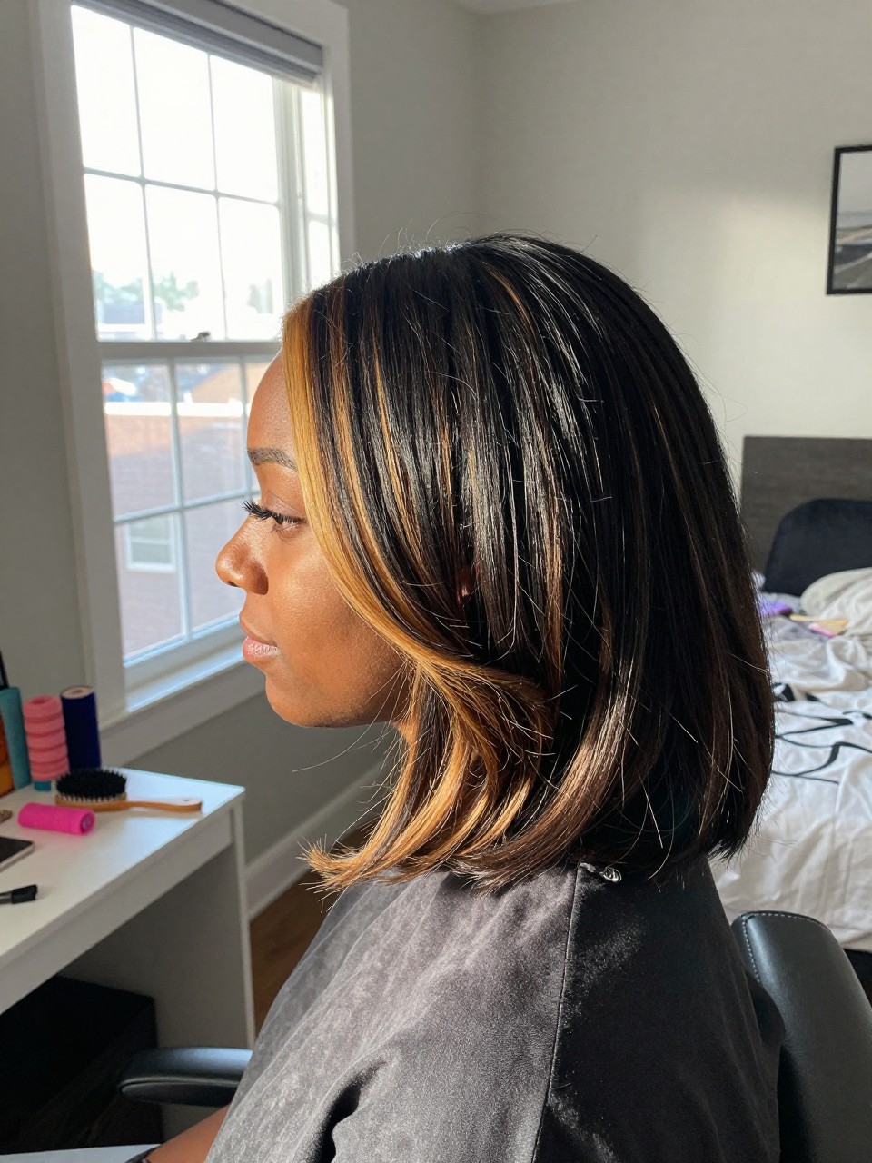 Photo of a black woman wearing a side part black blowout with warm blonde highlights, side profile view, in a sun-drenched bedroom, natural window light, containing a small lived-in detail such as velcro rollers on the vanity and a round brush nearby, latest iPhone photo quality.