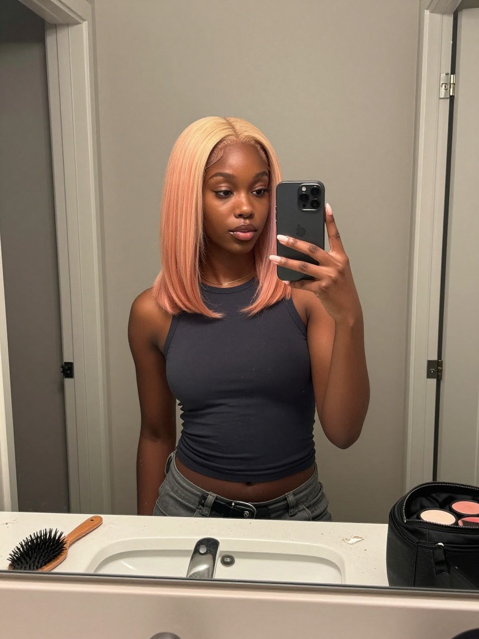Photo of a black woman wearing a sleek strawberry blonde lob with peachy blonde highlight ribbons, candid mirror selfie, at a bathroom vanity mirror, soft indoor lighting, containing a small lived-in detail such as a hairbrush on the vanity and a makeup bag nearby, latest iPhone photo quality.