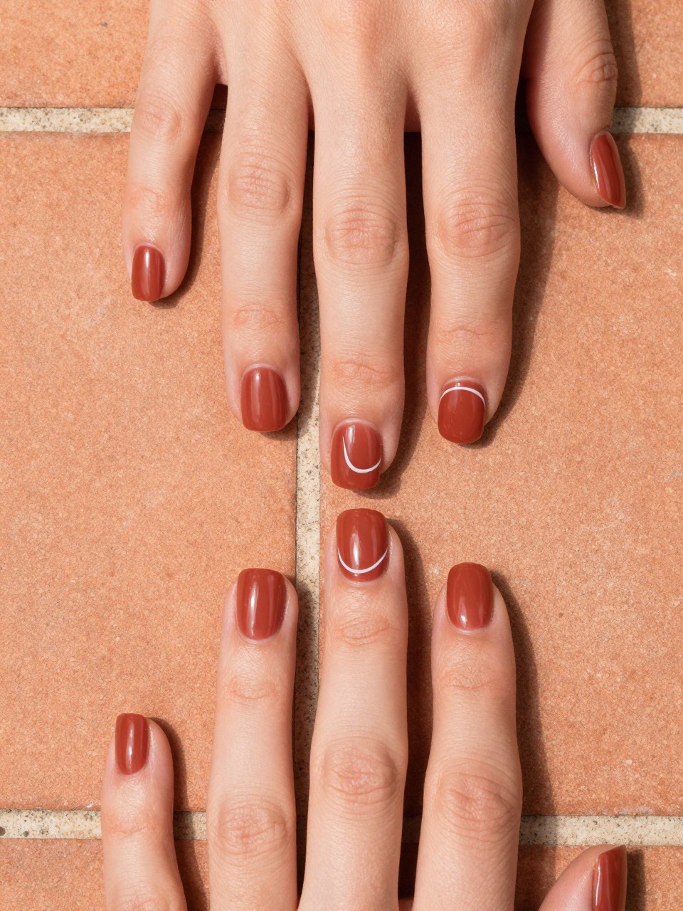 Image Prompt: Top-down view of hands with medium square nails on a terracotta tile. The nails are a warm, matte mulberry. A single, glossy clear curved line is painted on each nail, catching the light. The setting feels artistic and Mediterranean.
