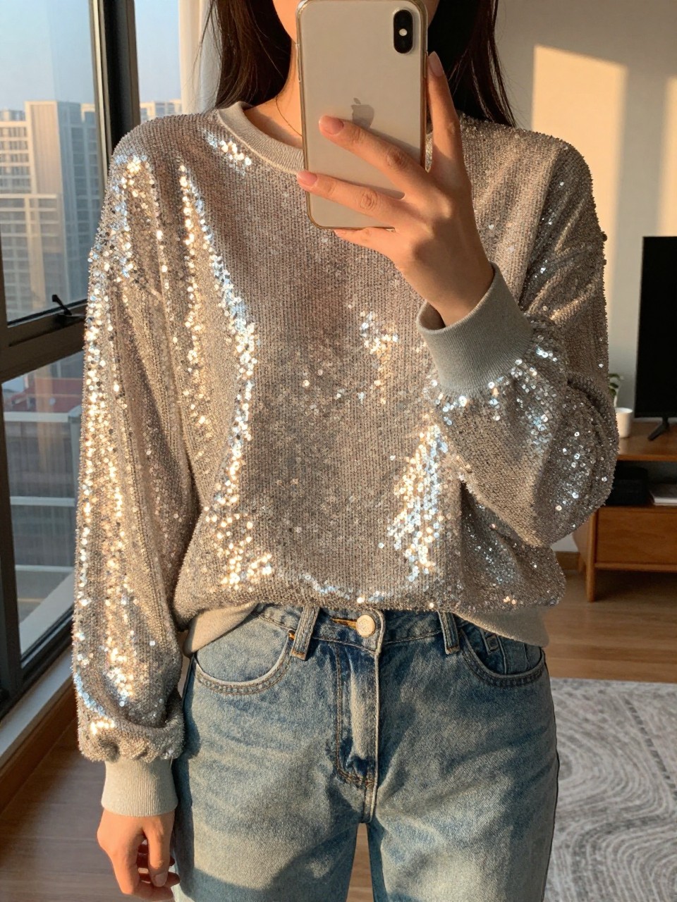 iPhone mirror selfie of a woman in a sequined sweatshirt and jeans, showing her phone case, cropped at NECK so face doesn't show, in a city apartment, evening golden hour, iPhone photo quality.