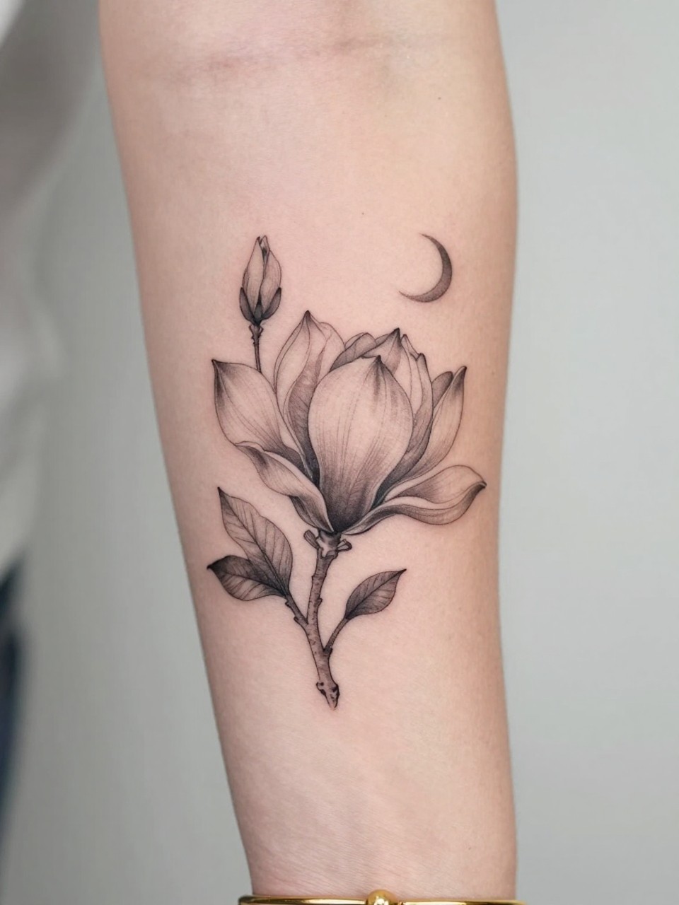 Image Prompt: "Design-focused close-up of a magnolia tattoo with a tiny crescent moon on the inner arm, black ink fine line floral design, broad petals, soft shading, delicate celestial accent, realistic skin texture, gold bracelet lightly visible, feminine modern tattoo inspiration photo, clean soft background, tattoo is the hero (no watermarks on images)"