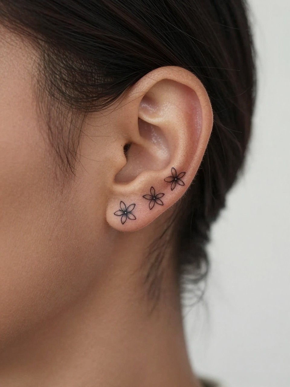 Image Prompt: "Macro shot of a tiny sampaguita flower tattoo behind the ear on dark skin, fine line black ink floral design with small star-shaped petals and soft shading, realistic skin texture, a few loose strands of hair framing the placement, minimal feminine styling, tight crop, soft clean background, tattoo-centered inspiration image (no watermarks on images)"