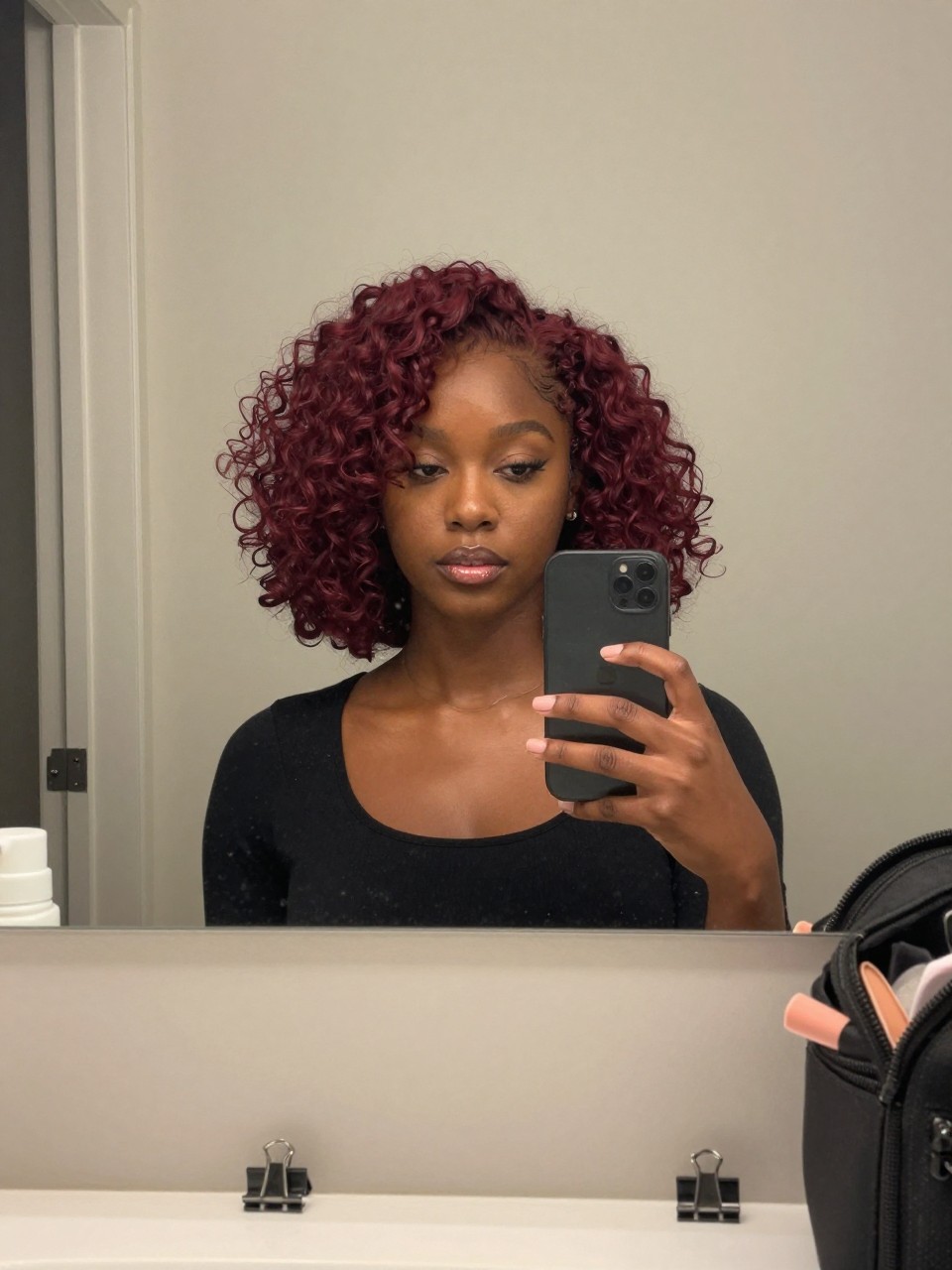 Photo of a black woman wearing a cherry red curly bob with soft defined curls, candid mirror selfie, bathroom vanity mirror setting, soft indoor lighting, with small claw clips on the counter and a makeup bag nearby, latest iPhone photo quality.