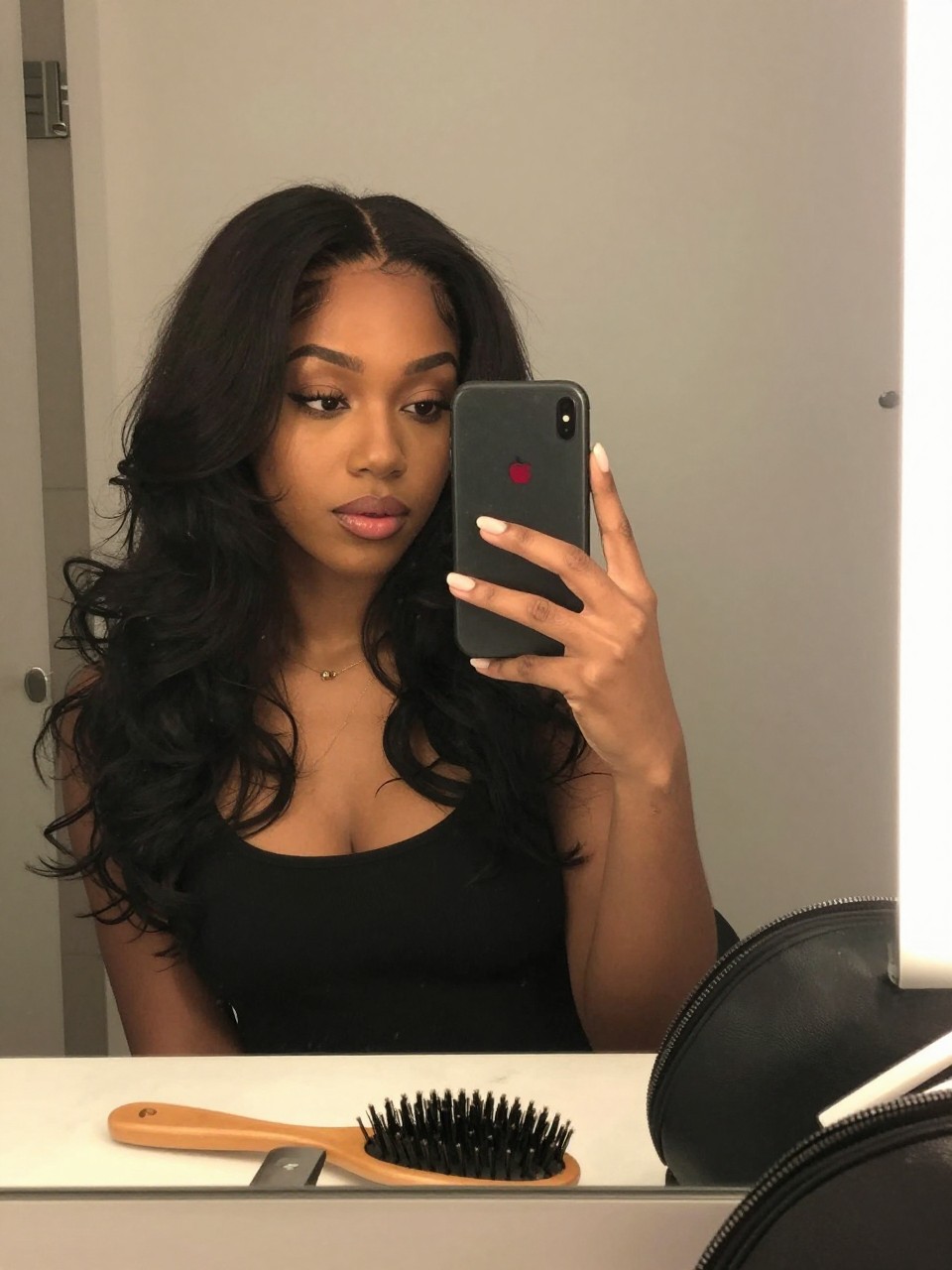 Photo of a black woman wearing dark hair with a cherry red money piece and soft face-framing waves, candid mirror selfie, bathroom vanity mirror setting, soft indoor lighting, with a hairbrush on the vanity and a makeup bag nearby, latest iPhone photo quality.