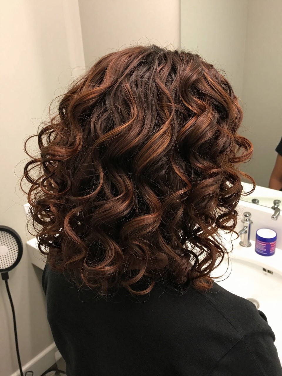 Photo of a black woman wearing cherry brown curls with soft highlights and defined volume, back-of-head view to highlight the highlighted curl pattern, bathroom vanity mirror setting, soft indoor lighting, with a diffuser on the counter and a curl cream jar nearby, latest iPhone photo quality.