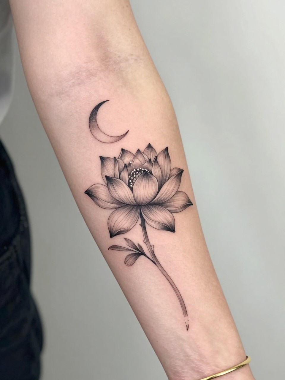 Image Prompt: "Design-focused close-up of a water lily tattoo with a tiny crescent moon on the inner arm, black ink fine line floral design, soft petal shading, delicate celestial accent, realistic skin texture, gold bracelet lightly visible, feminine modern tattoo inspiration photo, clean soft background, tattoo is the hero (no watermarks on images)"