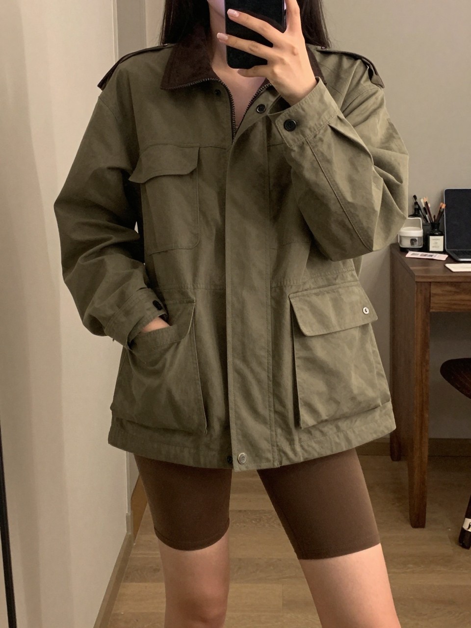 iPhone mirror selfie of a woman in a utility jacket and biker shorts, posing with hands in jacket pockets, cropped at NECK so face doesn't show, in a city apartment, warm moody lighting, iPhone photo quality.