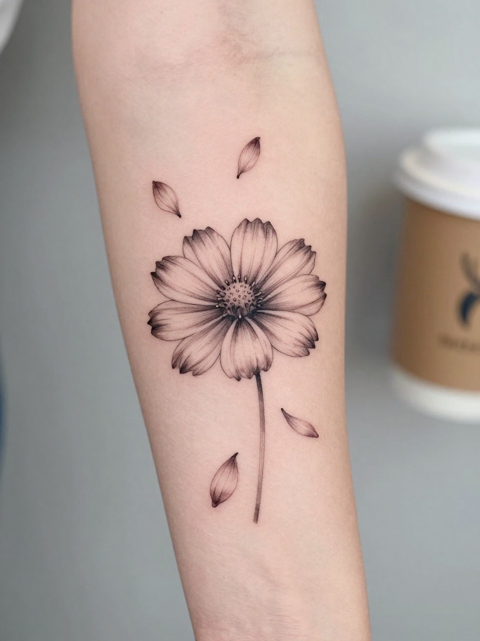 Extreme close-up of a cosmos tattoo with a few falling petals on the forearm, black ink fine line floral design, delicate petal movement, soft shading, realistic skin texture, coffee cup softly blurred nearby, modern feminine tattoo inspiration image, minimal background, tattoo-centered composition (no watermarks on images)