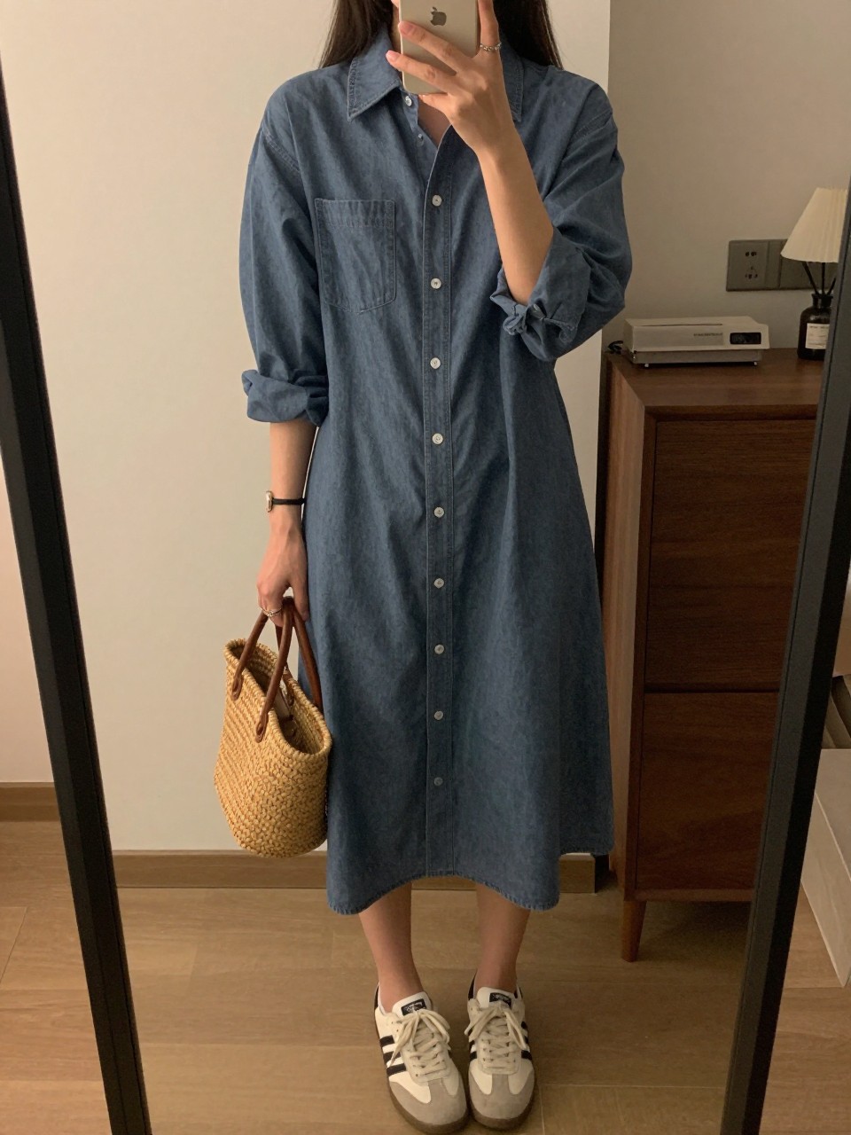 iPhone mirror selfie of a woman in a chambray shirtdress and sneakers, holding a basket bag, cropped at NECK so face doesn't show, in a city apartment, warm moody lighting, iPhone photo quality.