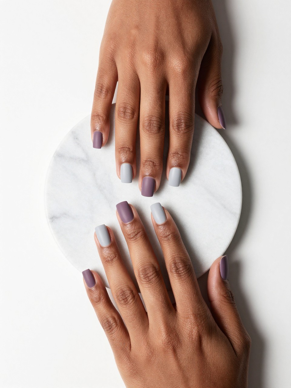 Image Prompt: Overhead shot of black feminine hands with short, squared nails placed on a modern marble coaster. The nails feature clean, diagonal color blocks of matte mauve and matte light grey. The background is minimalist and clean with bright lighting.