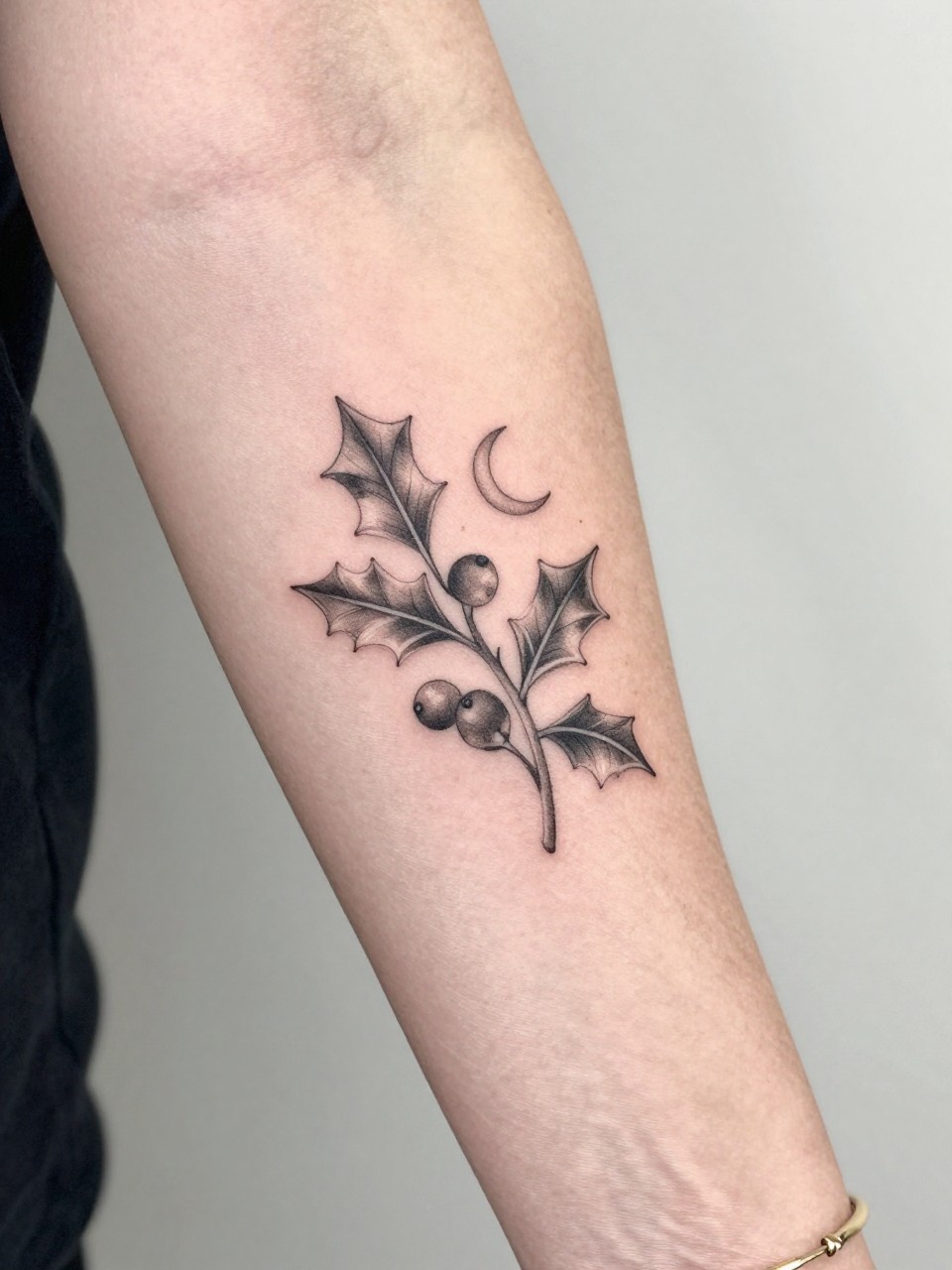 Image Prompt: "Design-focused close-up of a holly tattoo with a tiny crescent moon on the inner arm, black ink fine line leaves and berry details, soft shading, delicate celestial accent, realistic skin texture, gold bracelet lightly visible, feminine modern tattoo inspiration photo, clean soft background, tattoo is the hero (no watermarks on images)"