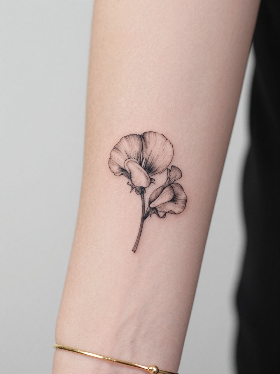 Image Prompt: "Macro shot of a tiny sweet pea flower tattoo on the inner wrist, black ink fine line floral design with soft curling petals and light shading, minimalist style, realistic skin texture, delicate gold bracelet, tight crop, feminine tattoo inspiration image, clean minimal background, tattoo-centered composition (no watermarks on images)"