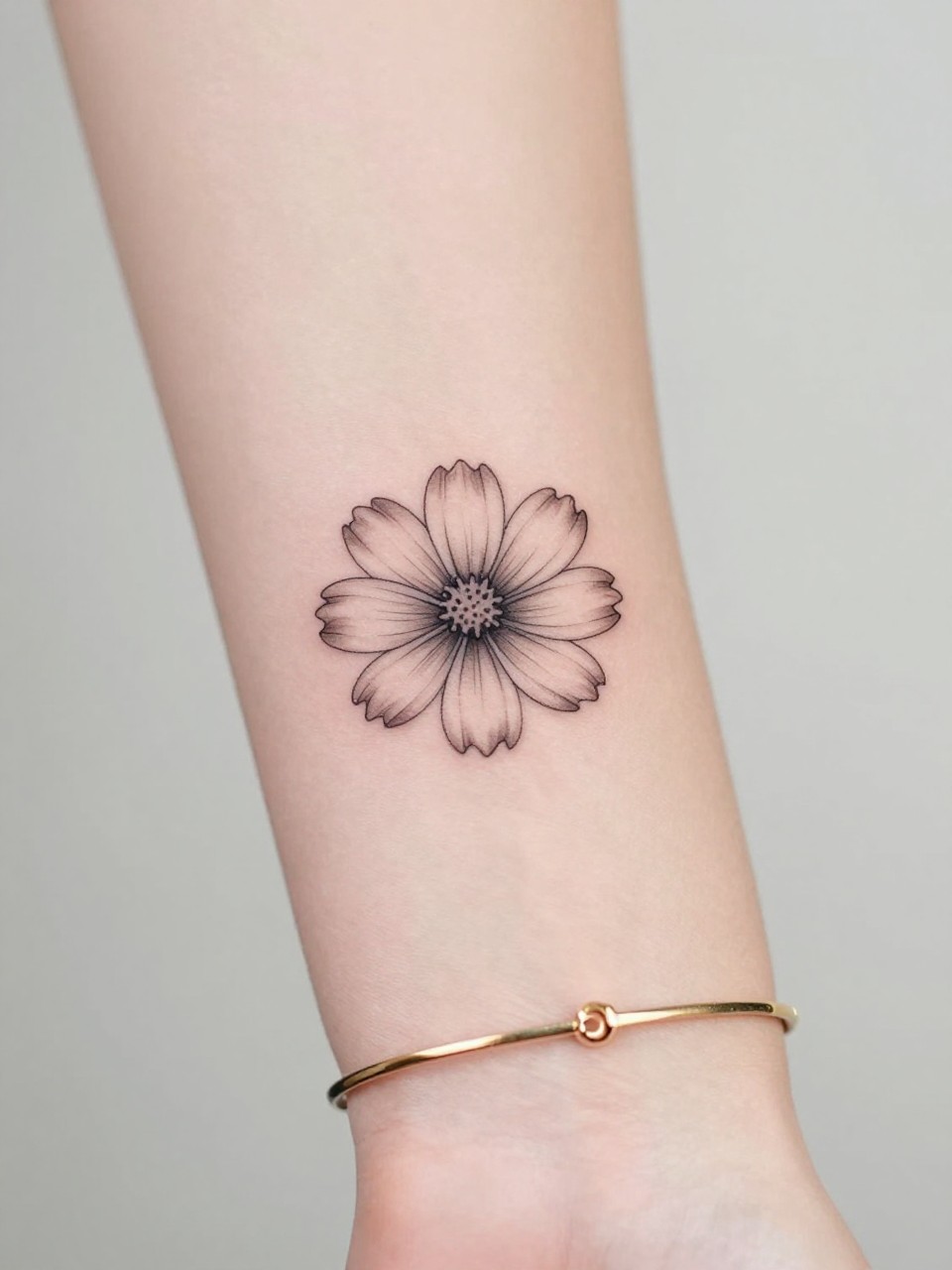 Macro shot of a tiny cosmos flower tattoo on the inner wrist, black ink fine line floral design with soft petal detail and light shading, minimalist style, realistic skin texture, delicate gold bracelet, tight crop, feminine tattoo inspiration image, clean minimal background, tattoo-centered composition (no watermarks on images)