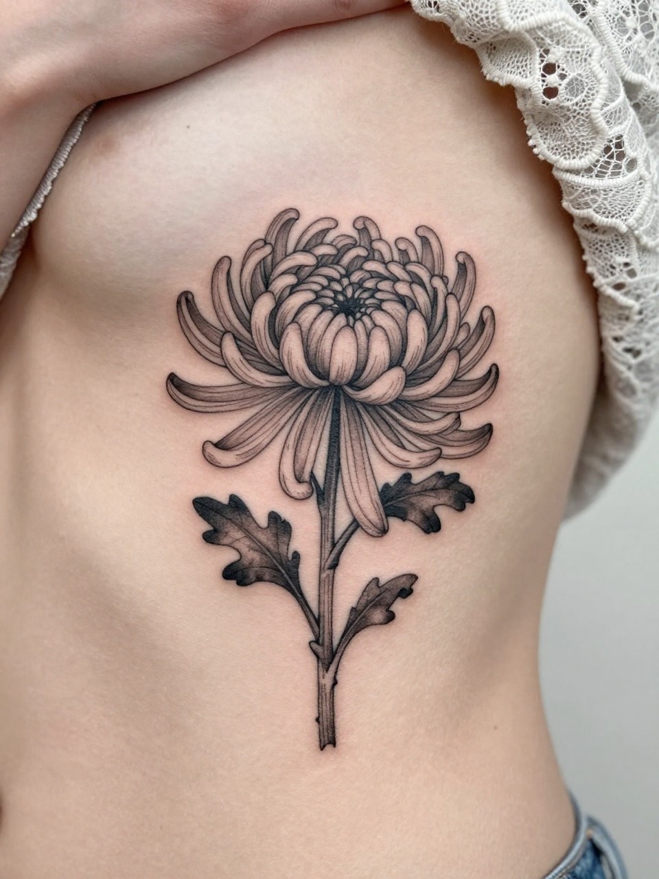 Image Prompt: "Design-focused close-up of a single chrysanthemum stem tattoo on the ribcage, black ink fine line floral design with many soft petals and light grey shading, realistic skin texture, lace fabric edge softly visible, feminine modern tattoo photo, minimal background, close-up tattoo visibility, tattoo dominates the frame (no watermarks on images)"