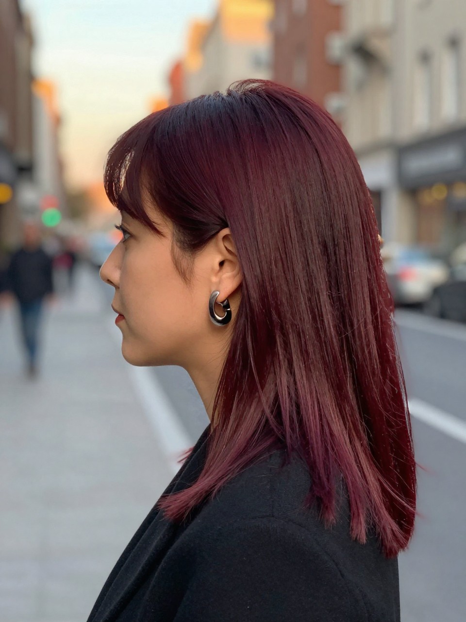 Photo of a woman wearing black cherry red hair styled sleek and straight, side profile view, softly blurred city street background, golden hour glow, with statement earrings peeking through and a hair clip clipped to her collar, latest iPhone photo quality.