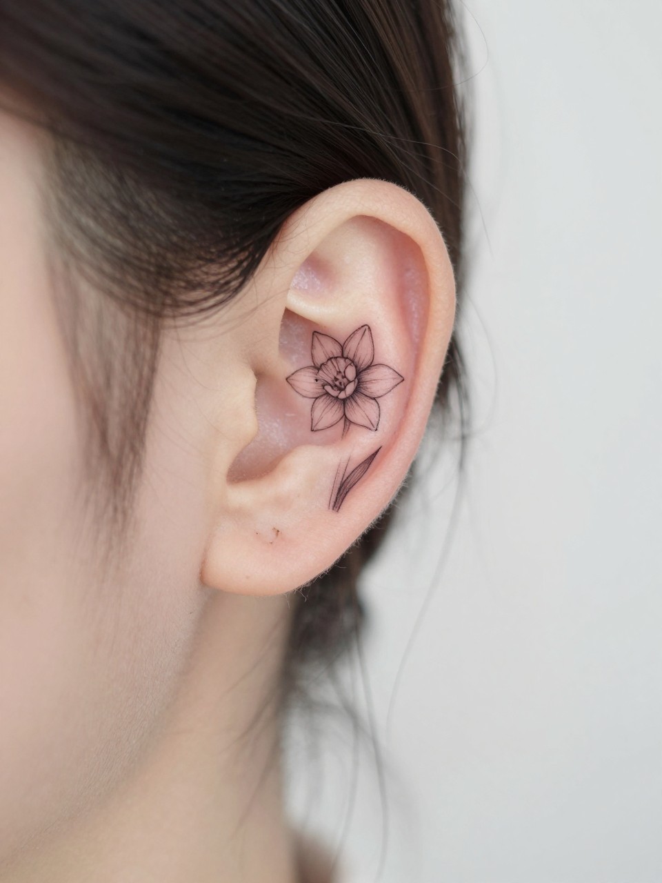 Image Prompt: "Macro shot of a tiny narcissus flower tattoo behind the ear, fine line black ink floral design with small delicate petals and soft shading, realistic skin texture, a few loose strands of hair framing the placement, minimal feminine styling, tight crop, soft clean background, tattoo-centered inspiration image (no watermarks on images)"