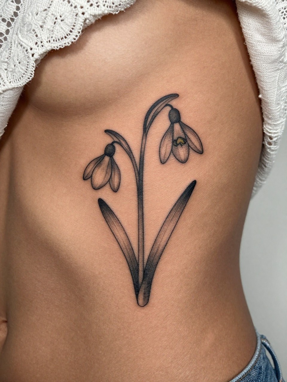 Image Prompt: "Design-focused close-up of a single snowdrop stem tattoo on the ribcage of a black woman, black ink fine line floral design with delicate drooping bloom and light grey shading, realistic skin texture, lace fabric edge softly visible, feminine modern tattoo photo, minimal background, close-up tattoo visibility, tattoo dominates the frame (no watermarks on images)"