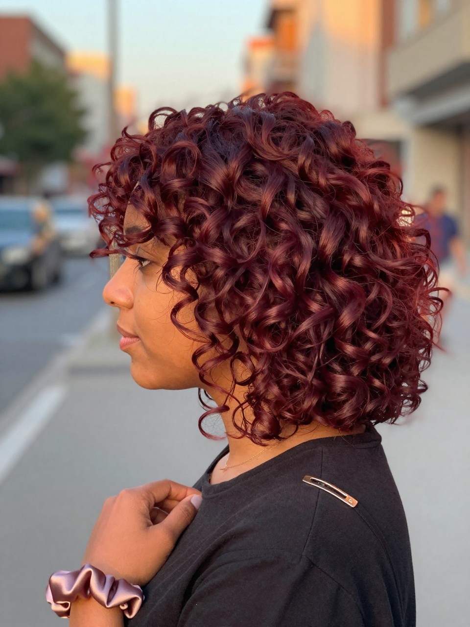 Photo of a brown woman wearing soft cherry red curls with defined volume, side profile view, softly blurred city street background, golden hour glow, with a silk scrunchie on her wrist and a hair clip clipped to her collar, latest iPhone photo quality.