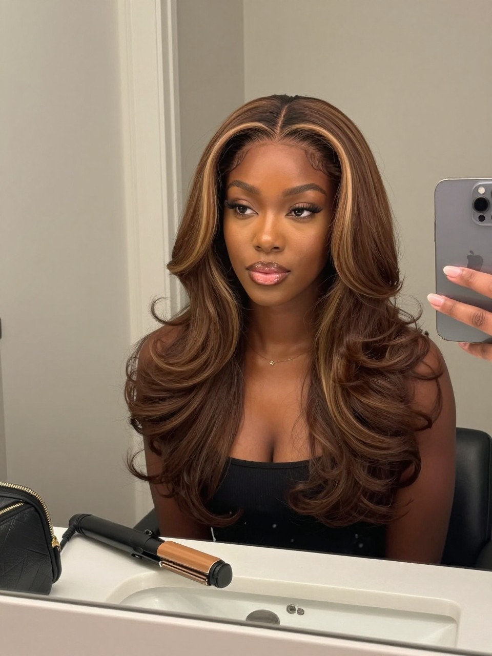 Photo of a black woman wearing mocha brunette hair with soft beige highlights styled in smooth loose bends, candid mirror selfie, bathroom vanity mirror setting, soft indoor lighting, with a flat iron on the counter and a small makeup bag nearby, latest iPhone photo quality.