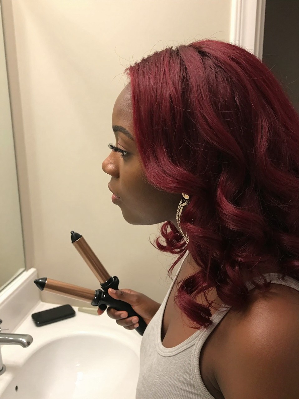Photo of a BLACK woman wearing ruby cherry red hair in soft loose curls, side profile view, bathroom vanity mirror setting, soft indoor lighting, with a curling iron on the counter and statement earrings peeking through, latest iPhone photo quality.