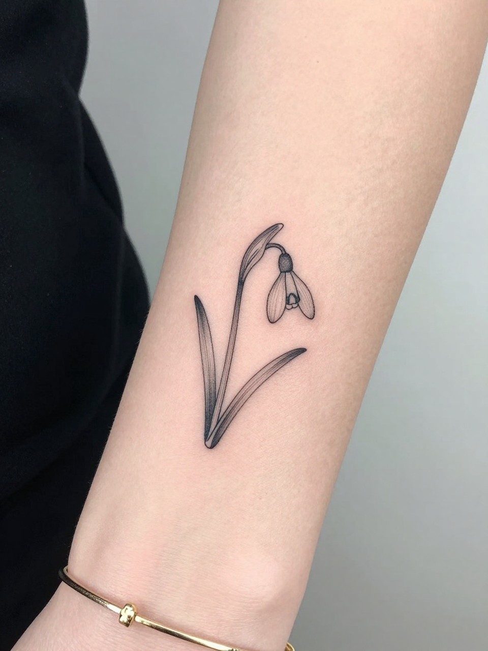 Image Prompt: "Macro shot of a tiny snowdrop flower tattoo on the inner wrist, black ink fine line floral design with a small drooping bloom and light shading, minimalist style, realistic skin texture, delicate gold bracelet, tight crop, feminine tattoo inspiration image, clean minimal background, tattoo-centered composition (no watermarks on images)"