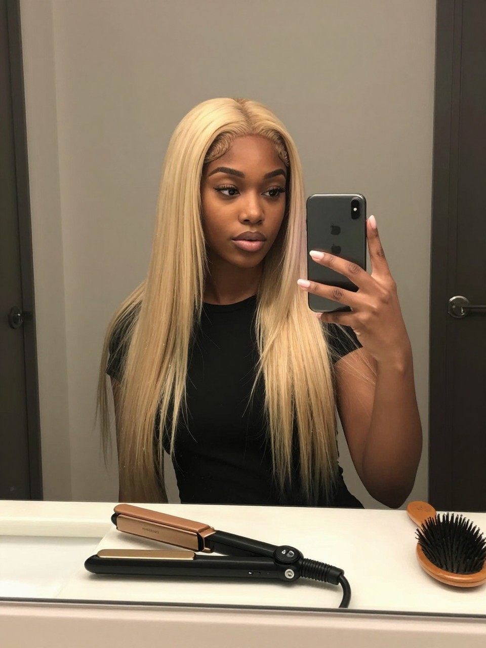 Photo of a black woman wearing icy blonde hair styled sleek and straight, candid mirror selfie, bathroom vanity mirror setting, soft indoor lighting, with a straightener on the counter and a hairbrush nearby, latest iPhone photo quality.