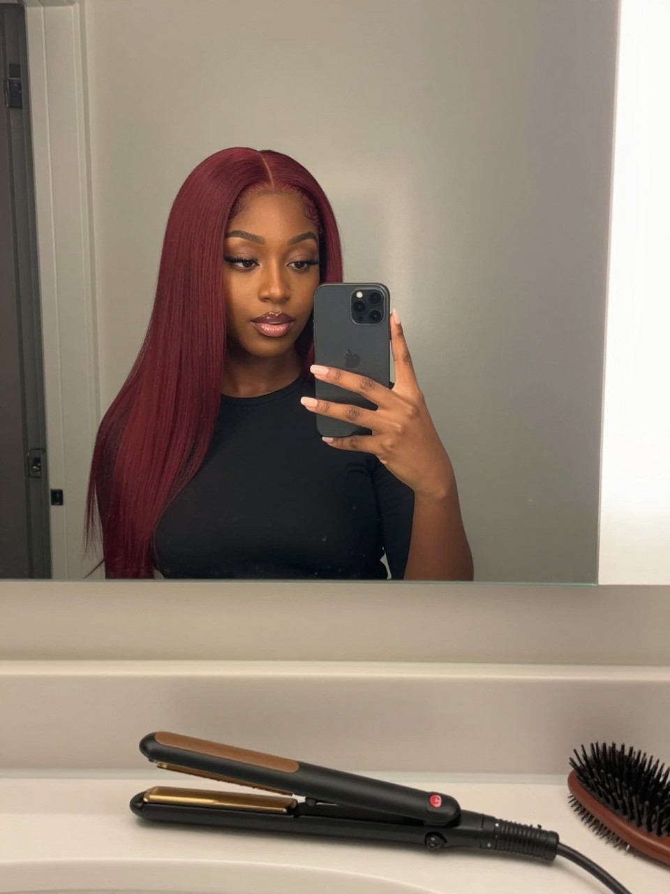 Photo of a black woman wearing candy cherry red hair styled sleek and straight, candid mirror selfie, bathroom vanity mirror setting, soft indoor lighting, with a straightener on the counter and a hairbrush nearby, latest iPhone photo quality.