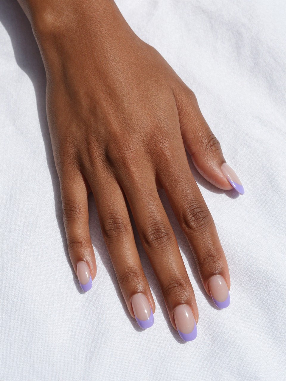 Image Prompt: Overhead shot of black feminine hands with long, natural oval nails resting on a white linen sheet. The nails have a clear, glossy base with crisp, thin tips painted in a soft matte lilac. The sunlight is bright and clean, highlighting the delicate color contrast.