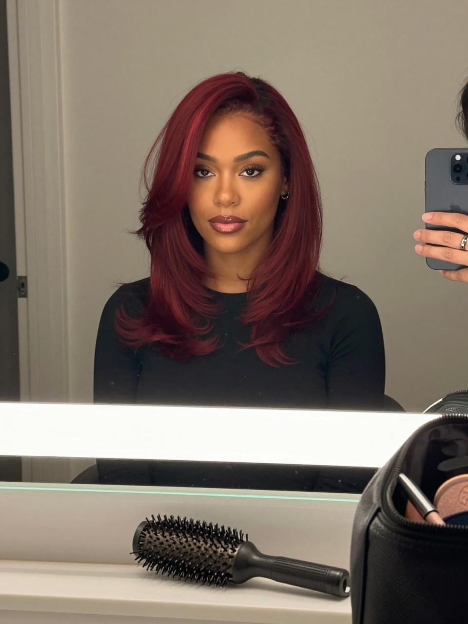Photo of a brown woman wearing cherry red hair with burgundy lowlights in a smooth layered blowout, candid mirror selfie, bathroom vanity mirror setting, soft indoor lighting, with a blowout brush on the counter and a makeup bag nearby, latest iPhone photo quality.